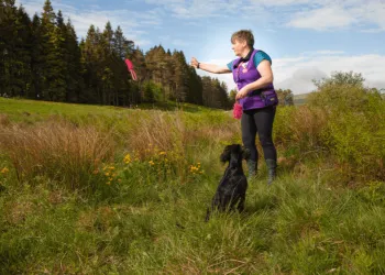 Woman with short hair and a purple top walks through stunning countryside carrying a pink dog lead