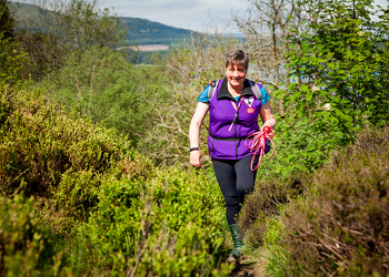 Woman with short hair and a purple top walks through stunning countryside carrying a pink dog lead