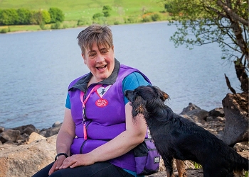 Woman with short hair and a purple top sits looking at a small black and tan spaniel who is off lead and is looking back