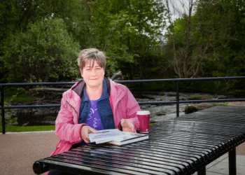 Woman in a pink jacket sits with a coffee and a pile of books next to a river.