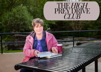 Image text reads The High Prey Drive Club. Woman with short hair and a pink jacket sits an outdoor table with a pile of dog training books and a coffee. A beautiful river flows behind her.